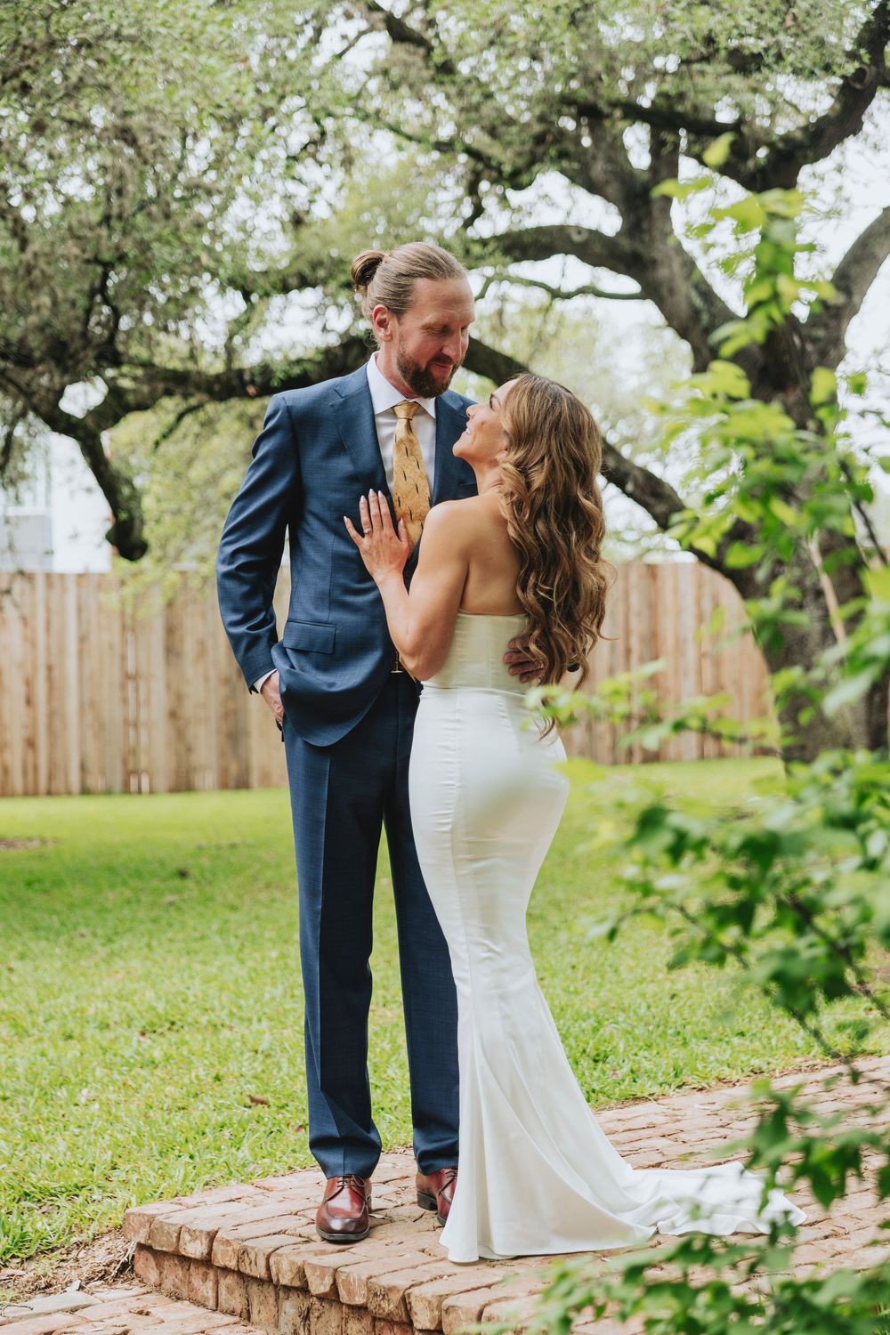 Bride and groom share a romantic moment outdoors, she in a white gown, he in a navy suit, under a large oak tree.