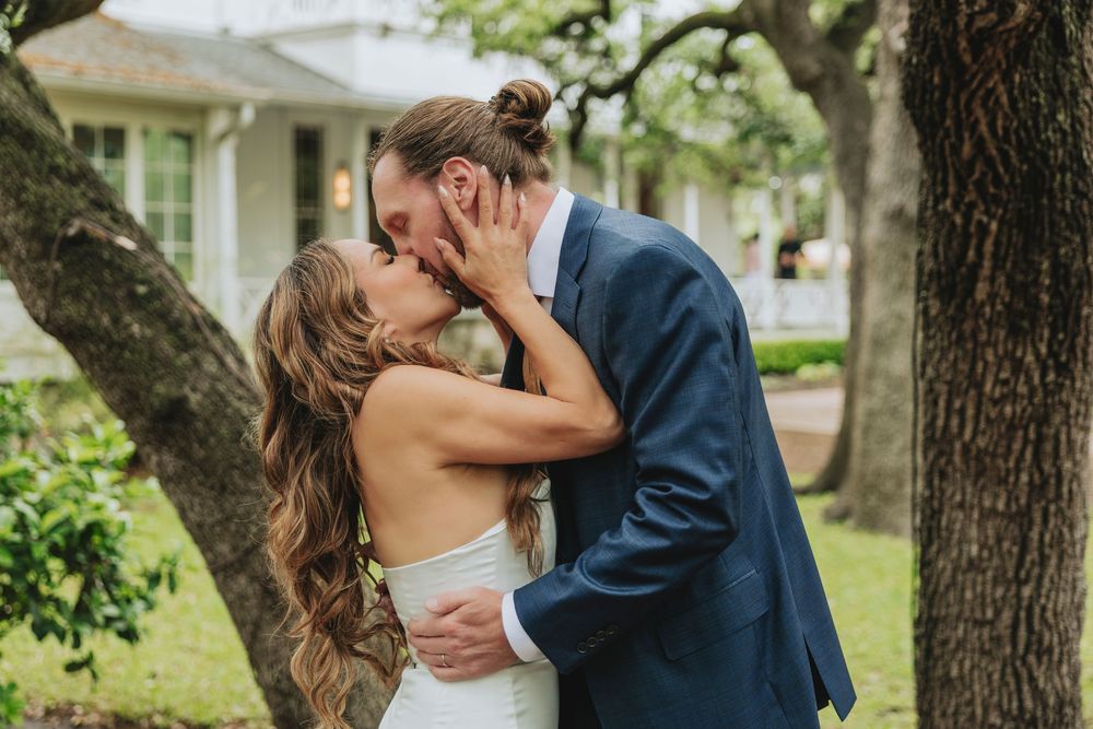 Bride and groom share a romantic kiss outdoors under oak trees, bride in white gown, groom in navy suit.