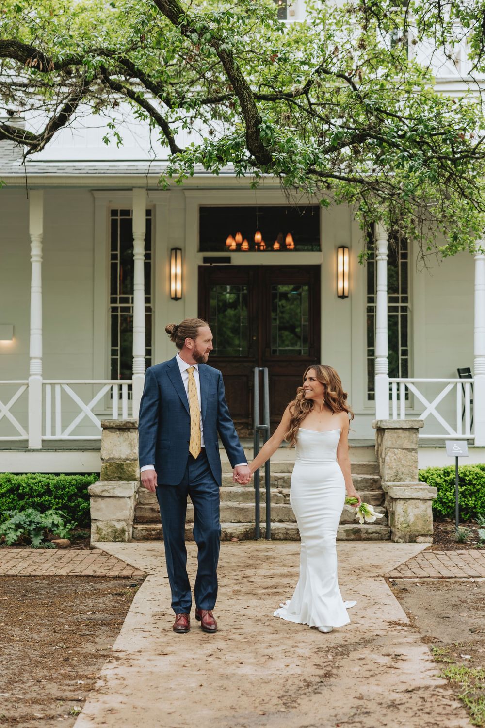 Bride and groom holding hands walking in front of elegant white venue with stone steps and lush trees.
