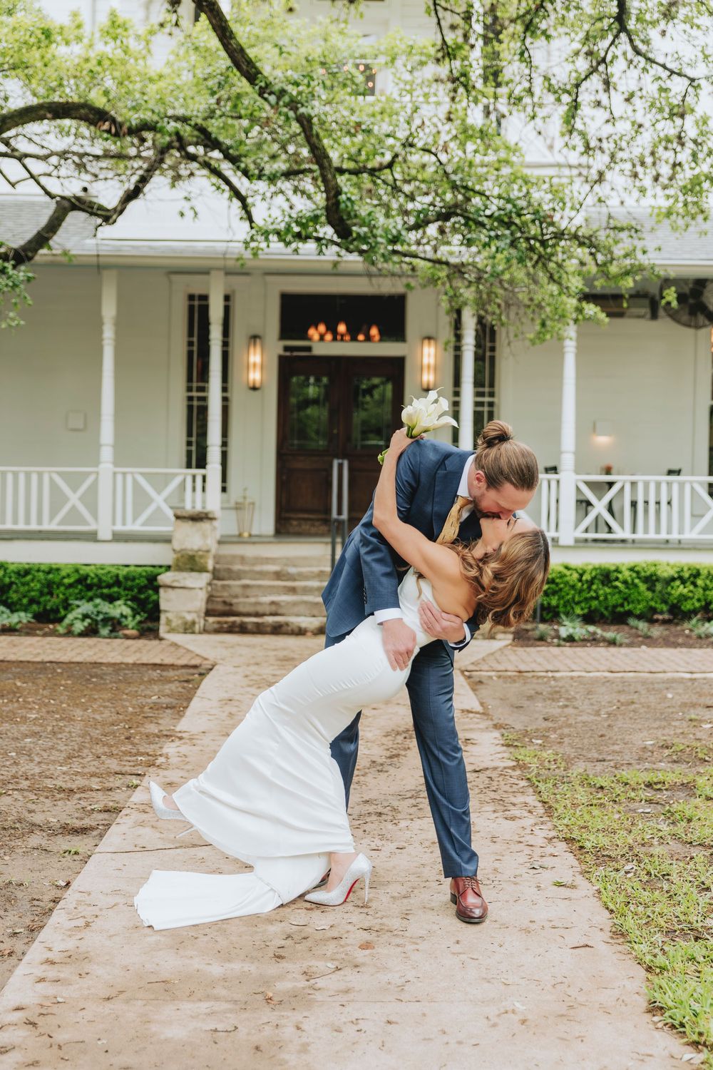 Groom dips bride for a romantic kiss on a garden path in front of a white Southern-style wedding venue.