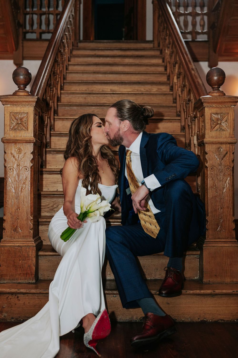 Bride and groom sharing a romantic kiss on ornate wooden staircase, she in white gown holding calla lilies, he in navy suit.