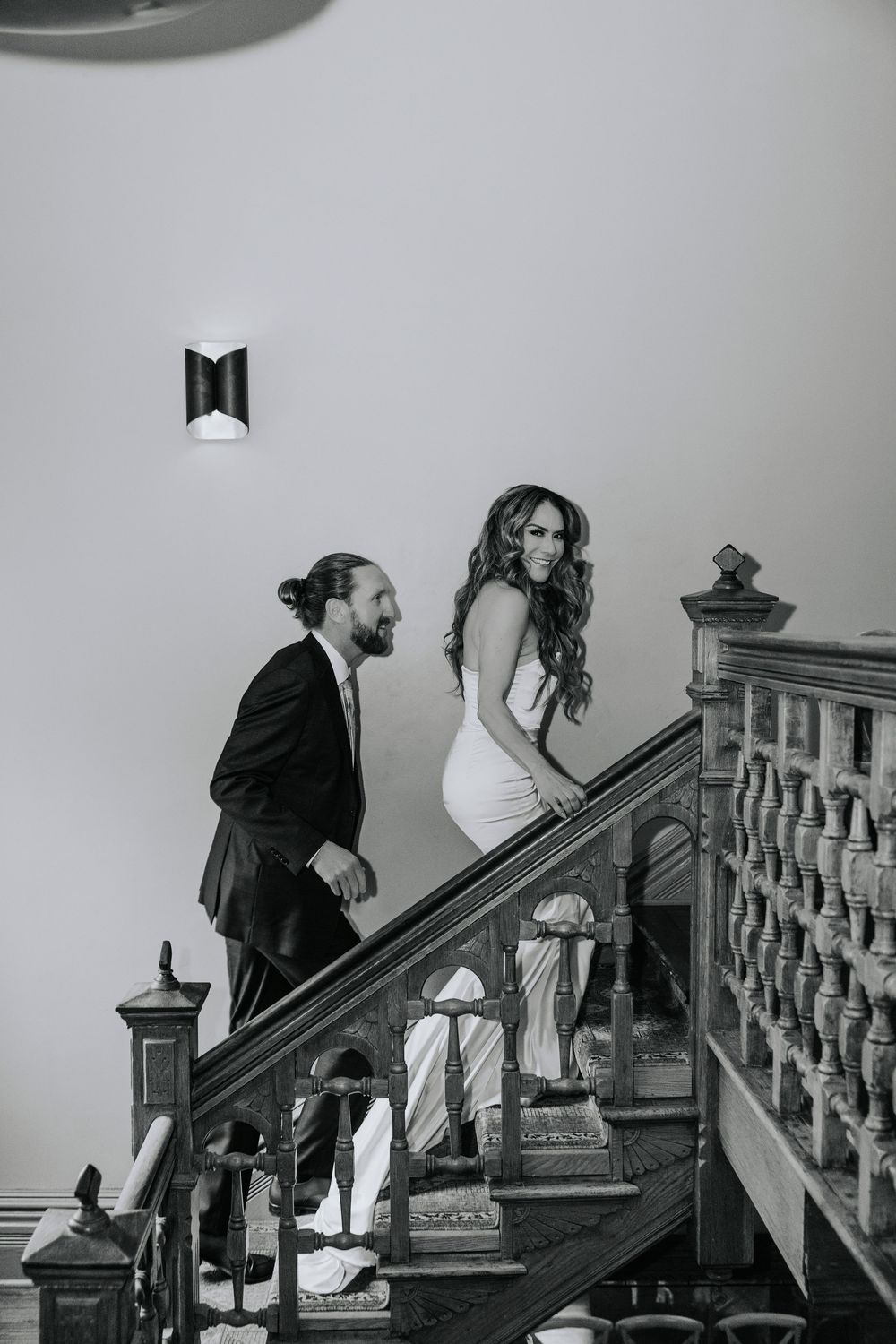 Black and white photo of a couple in formal attire walking up an elegant staircase, woman in white dress smiling back.