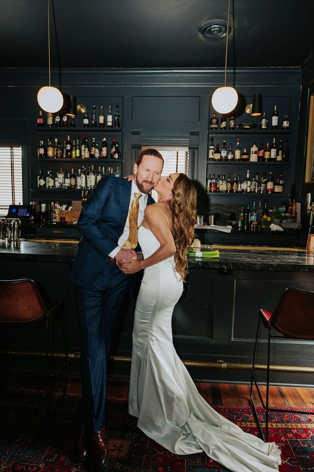 Bride and groom share a romantic kiss at a dark, moody bar backdrop on their wedding day.