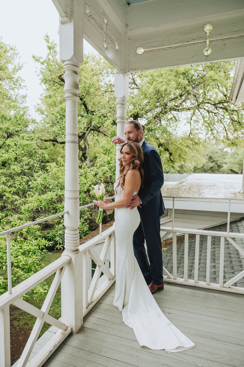 Bride in white gown and groom in navy suit pose on a white Victorian porch surrounded by lush green trees.