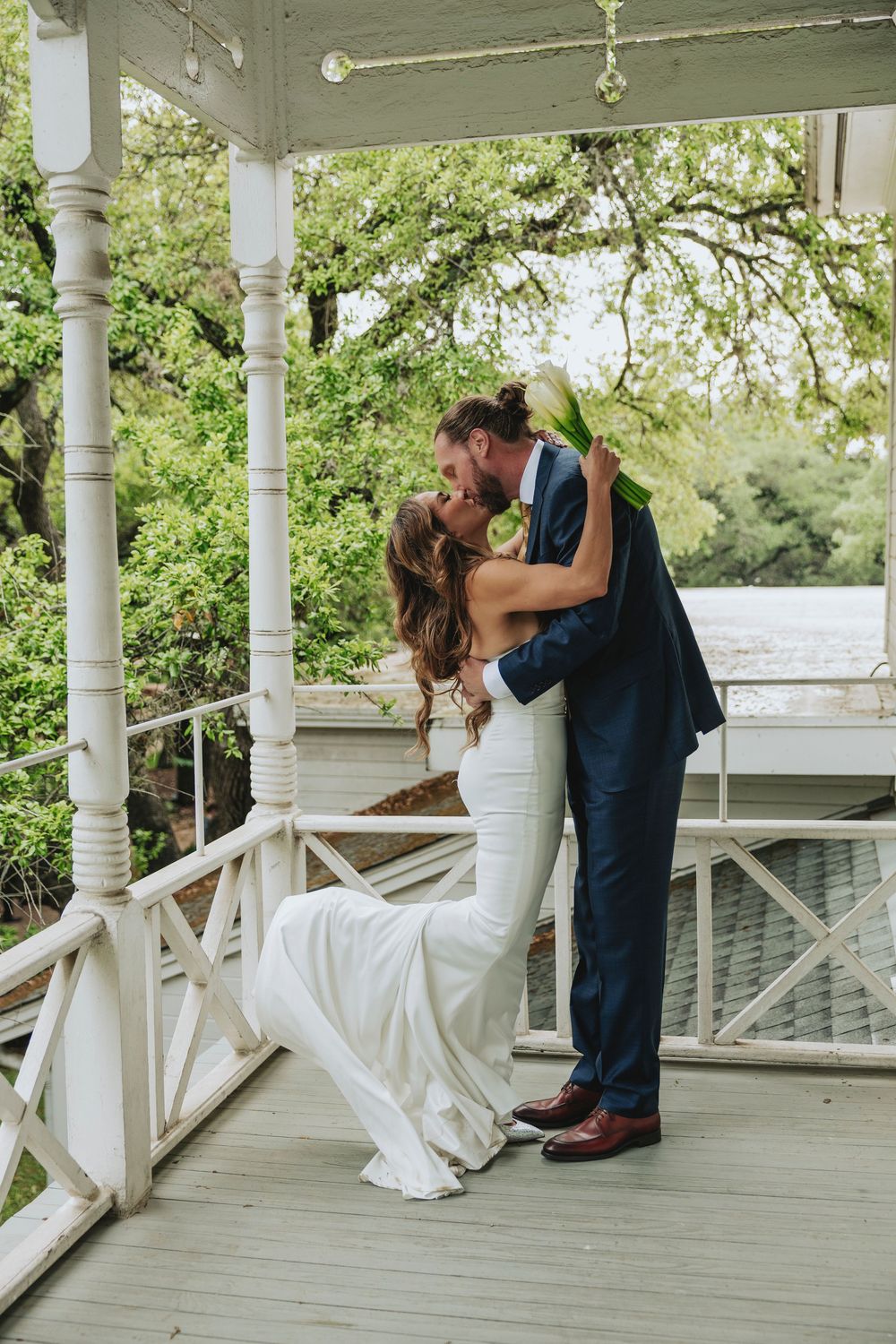 Bride and groom sharing a romantic kiss on a white gazebo porch, bride in fitted white gown holding bouquet.