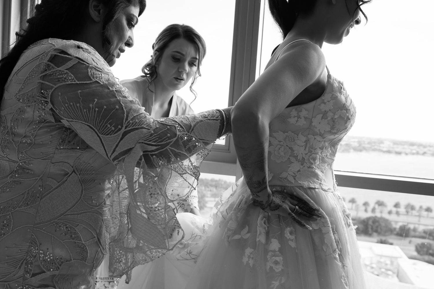 Black and white photo of bride being helped into wedding gown by two women near a large window with scenic view.
