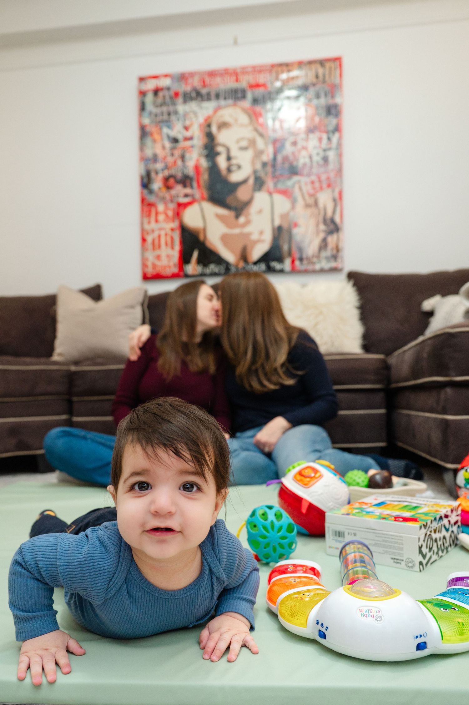 Baby doing tummy time on green mat with toys while two women sit on brown couch in living room.