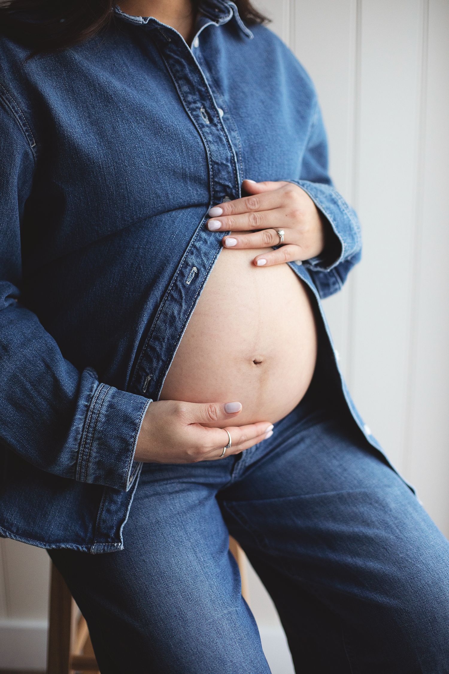 Pregnant woman in denim shirt and jeans cradling her baby bump with both hands, wearing rings. During a South east London maternity session.