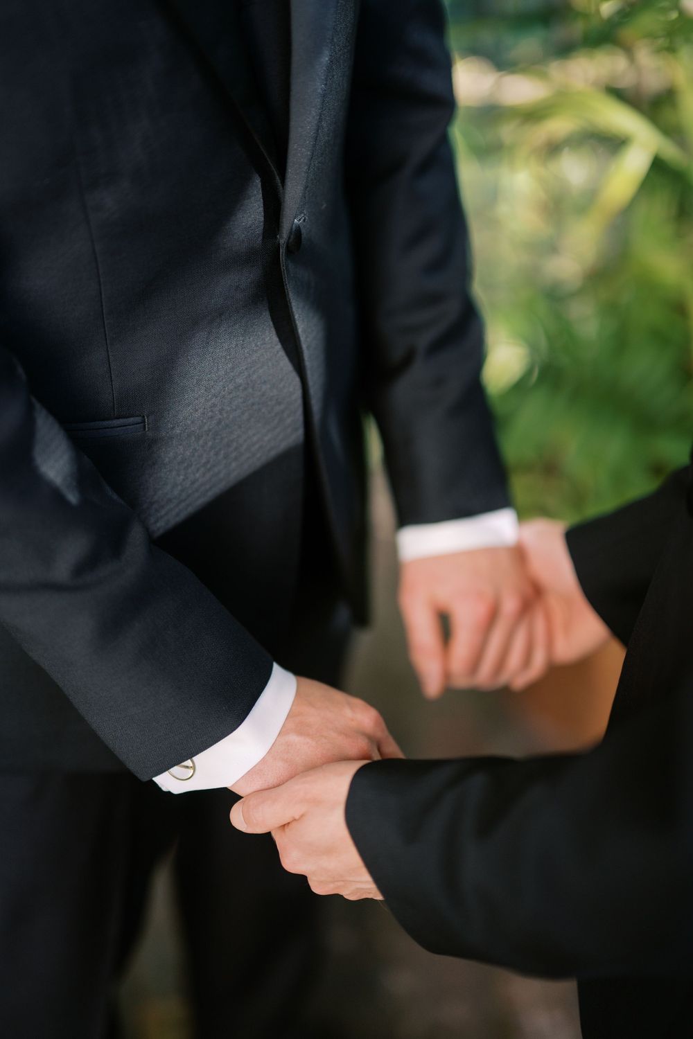 Close-up of two grooms in black suits holding hands during their wedding ceremony.