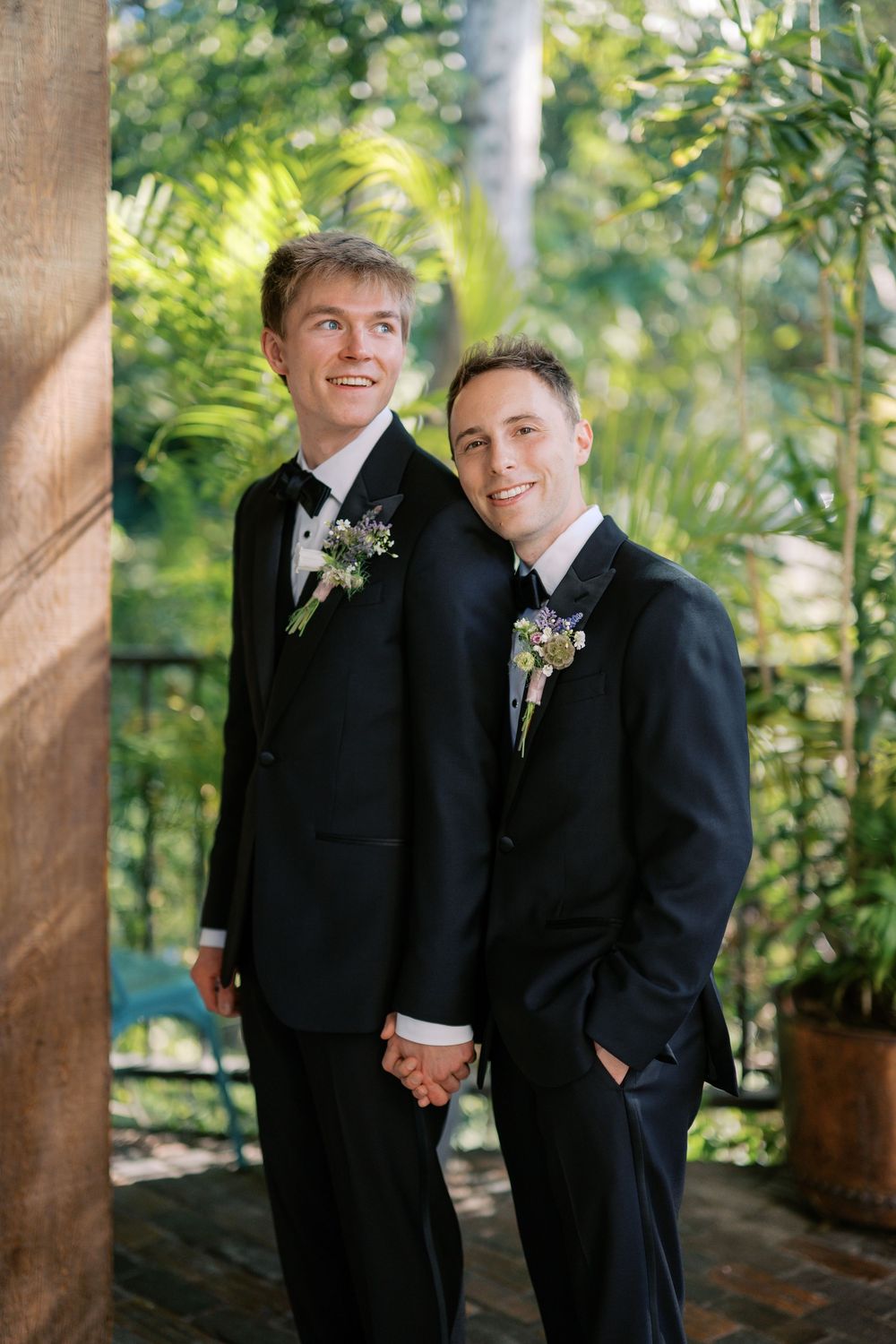 Two grooms in black tuxedos hold hands and smile among lush tropical greenery at their wedding.