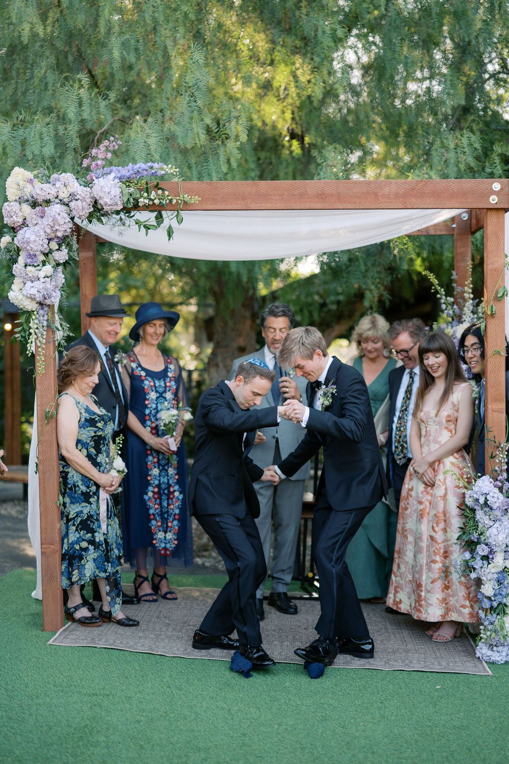 Two grooms share a joyful moment under a floral wedding arch surrounded by smiling guests outdoors.