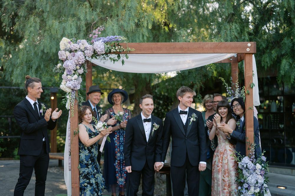Two grooms hold hands under a floral wooden arch, surrounded by applauding guests at an outdoor wedding ceremony.