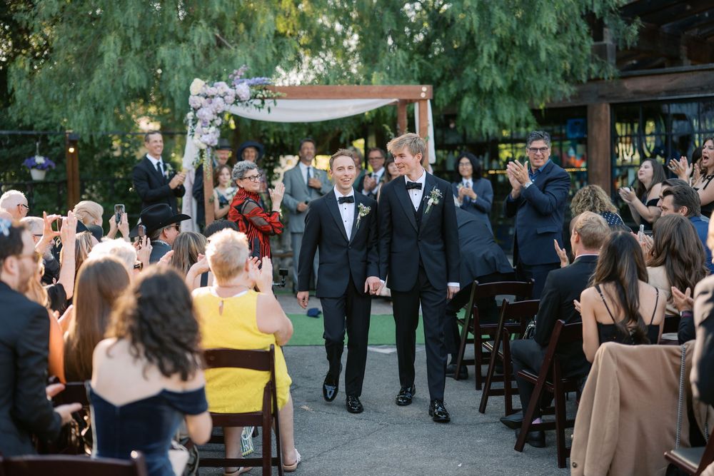 Two grooms in tuxedos walk hand-in-hand down the aisle at an outdoor wedding ceremony.