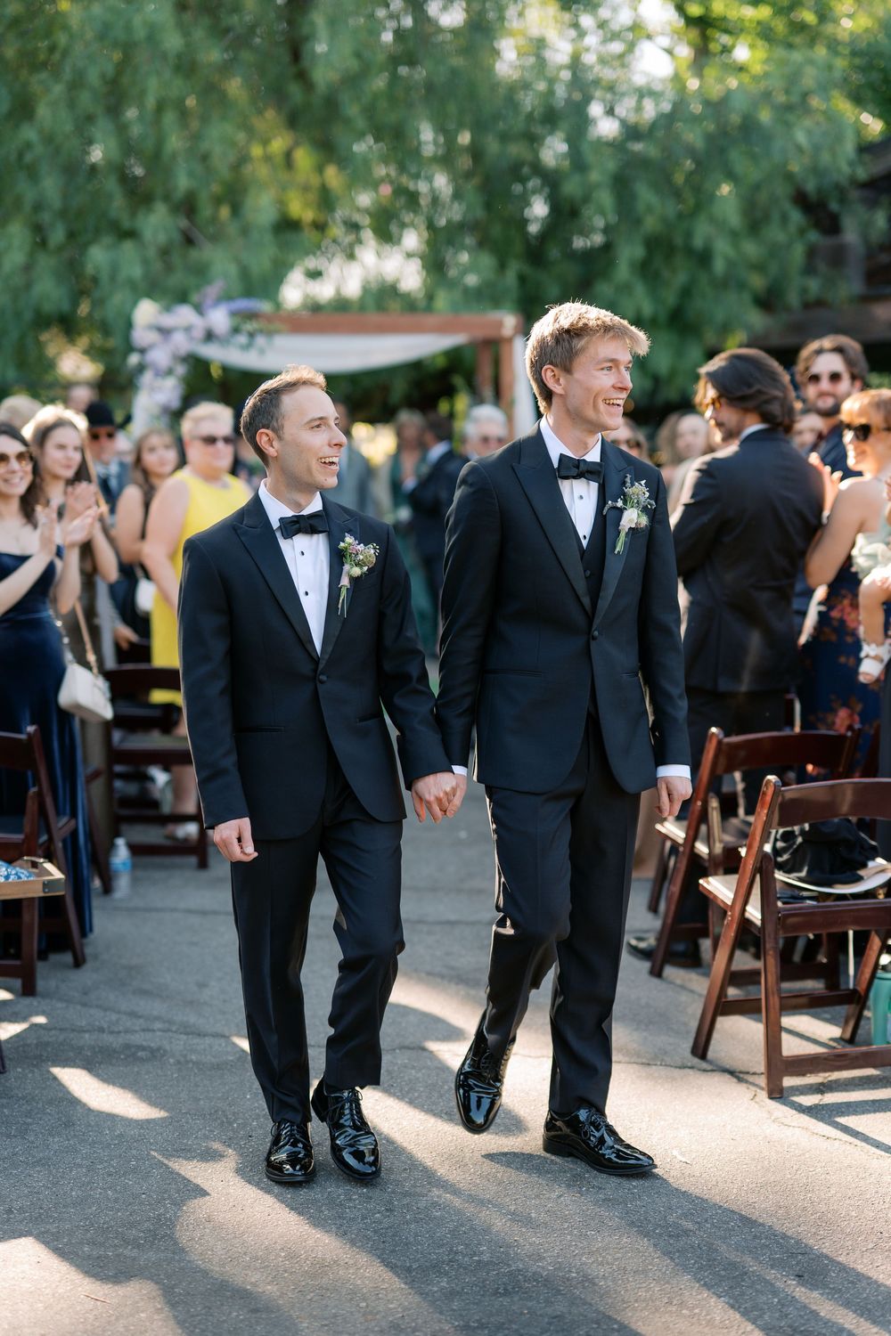Smiling grooms in matching black tuxedos walk hand in hand down sunlit outdoor wedding aisle.