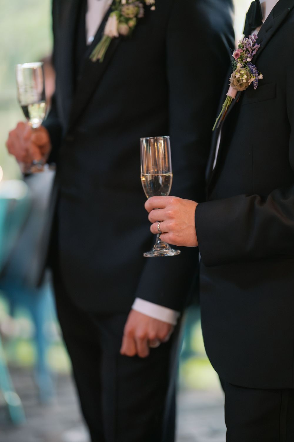 Two grooms in black tuxedos hold champagne flutes with wedding ring visible during reception.