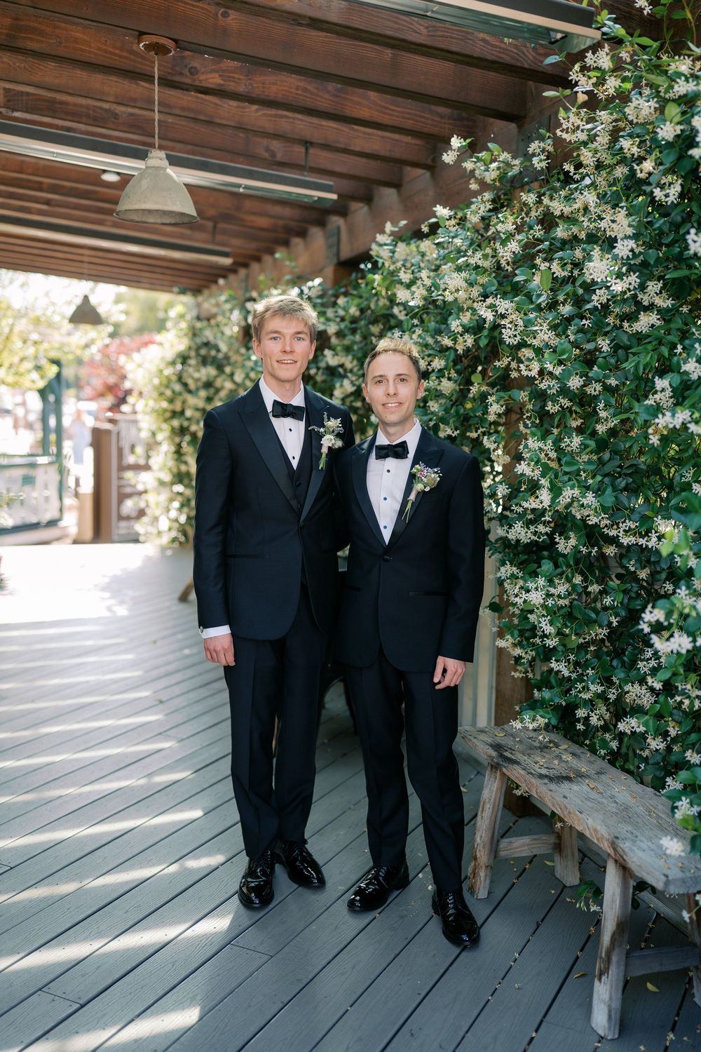 Two grooms in matching black tuxedos pose together under a pergola covered in white flowering vines.