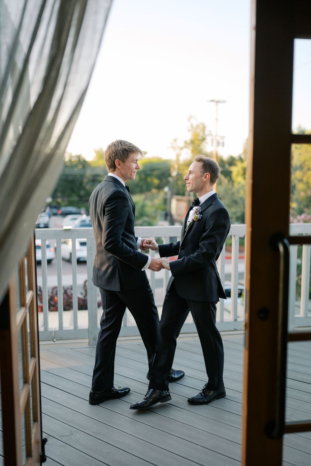 Two grooms in black suits share a tender moment on a wooden deck during their wedding celebration.