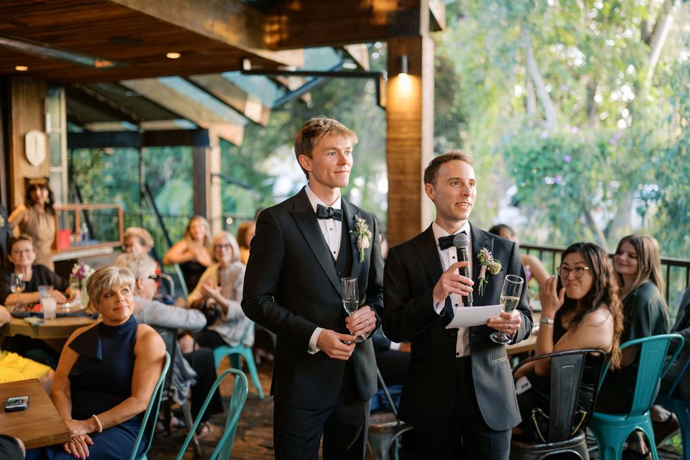 Two grooms in black tuxedos address wedding guests during an outdoor reception speech.