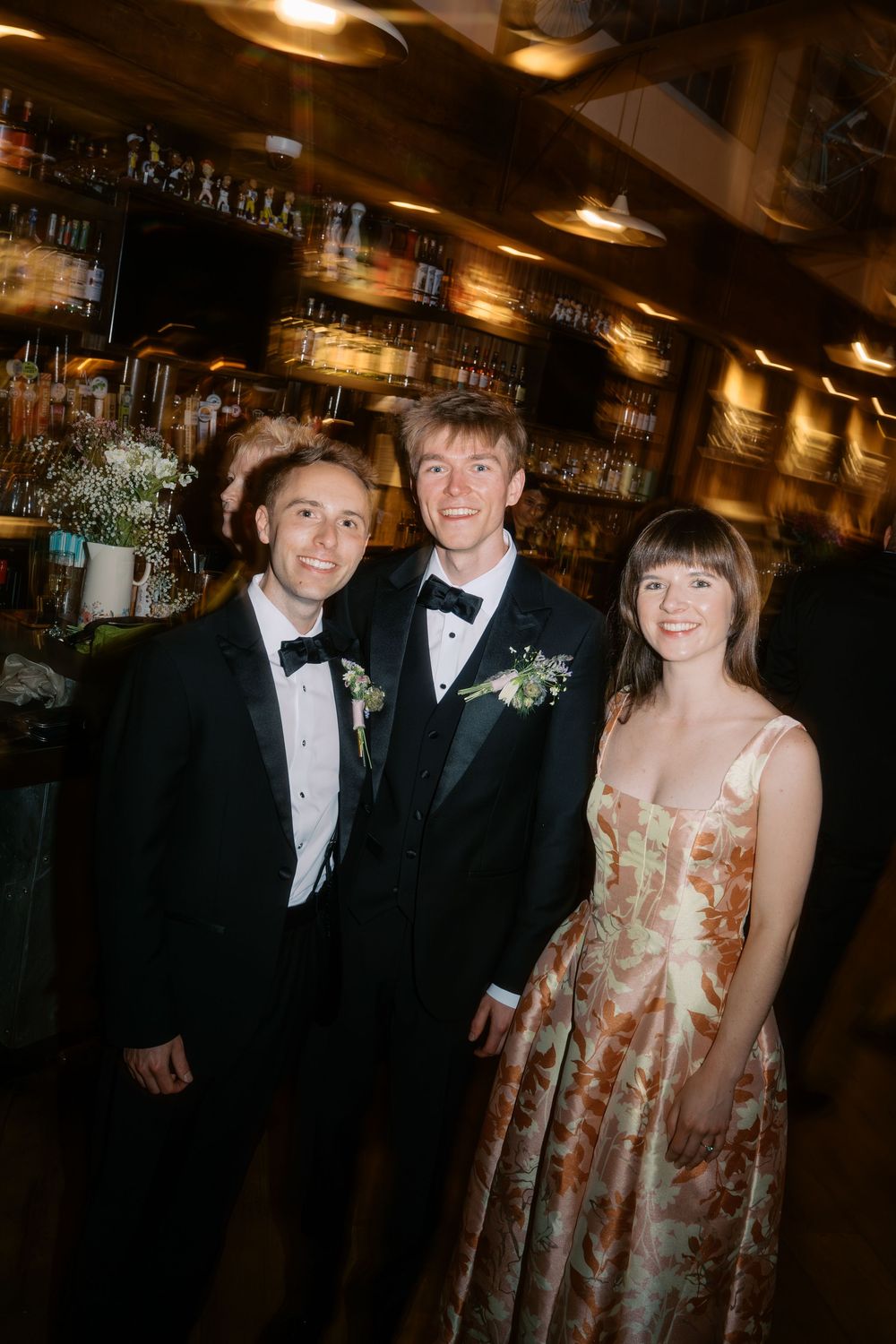 Two grooms in black tuxedos pose with a female wedding guest at a warmly lit reception bar.