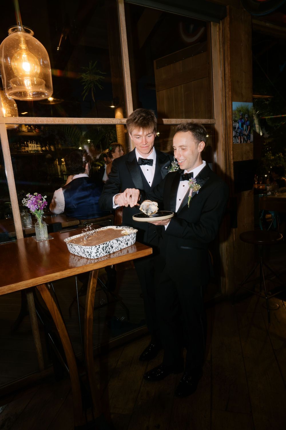 Two grooms in tuxedos cut their wedding cake together at a rustic indoor reception venue.