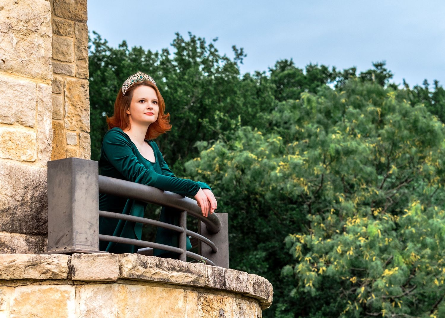 Red-haired woman in green dress poses on stone balcony overlooking lush green trees at golden hour.