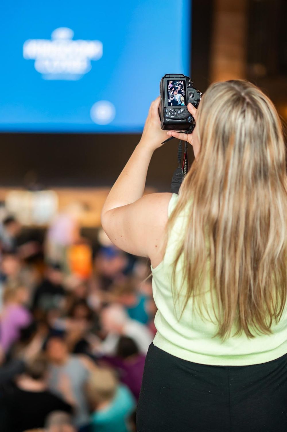 A blonde woman in a green tank top photographs an event crowd with a digital camera from behind.