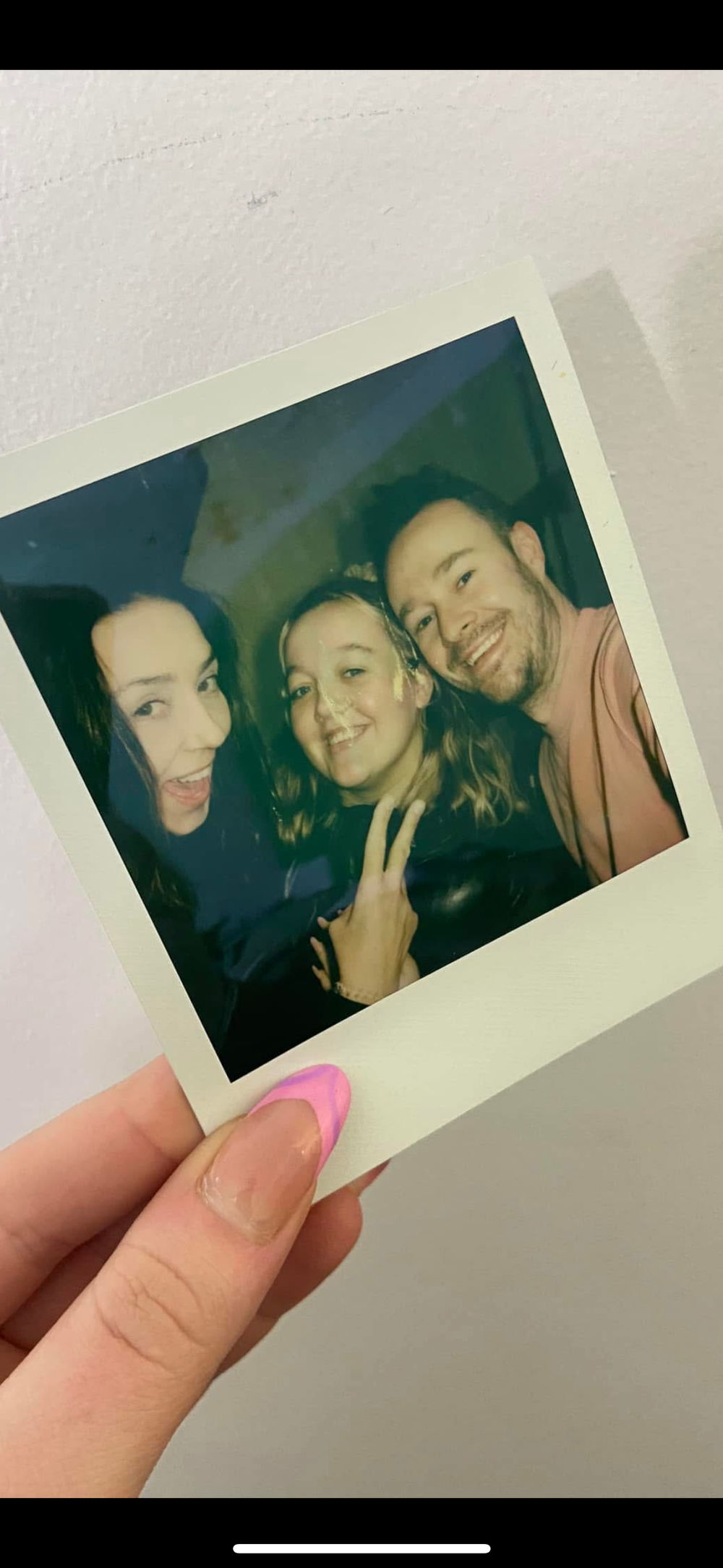Three friends posing together in a Polaroid photo, one making a peace sign, held by a hand with pink nails.