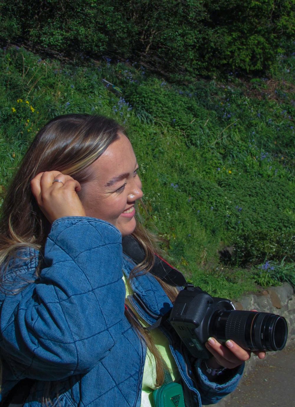 Woman with long hair smiling while holding a DSLR camera outdoors with green foliage in the background.