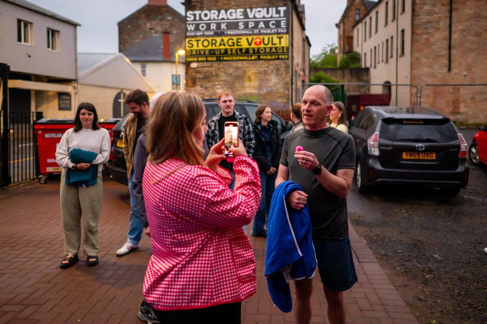 A woman photographs a man speaking outside a Storage Vault workspace on an overcast day.