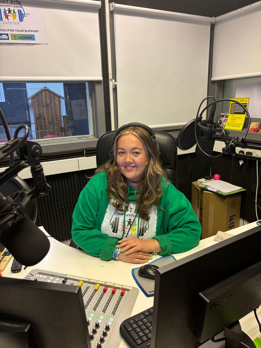 Woman in green sweatshirt sitting at radio studio desk with microphone.