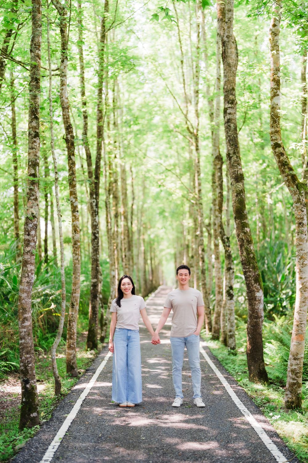 Couple holding hands walking on a tree-lined path in a lush green forest, wearing casual beige outfits.