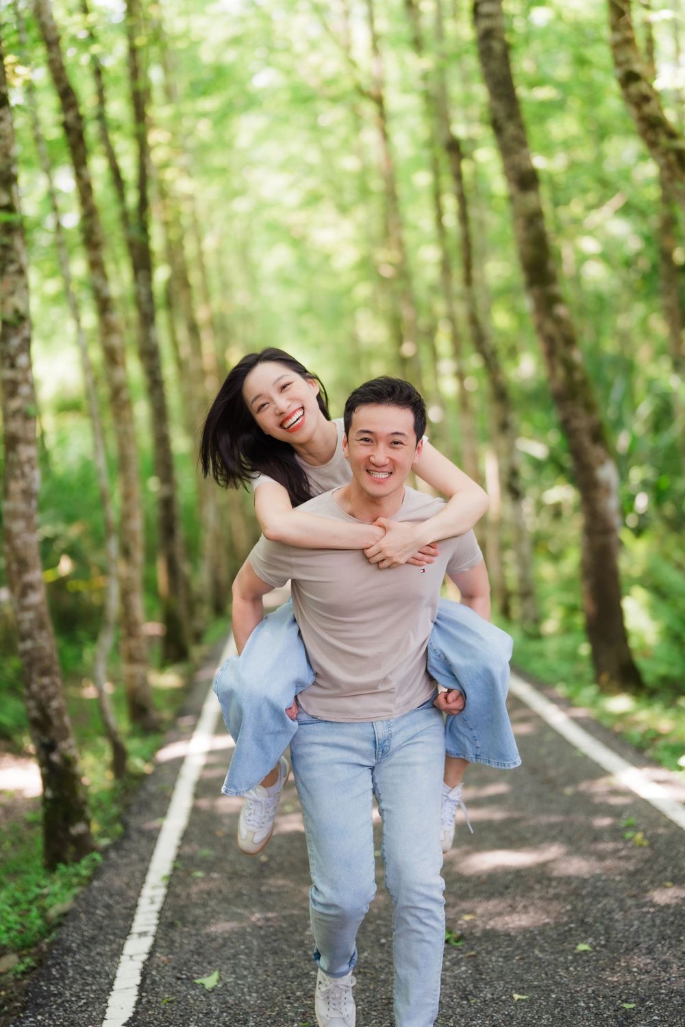 Happy Asian couple enjoying a piggyback ride on a scenic tree-lined path in a lush green forest.