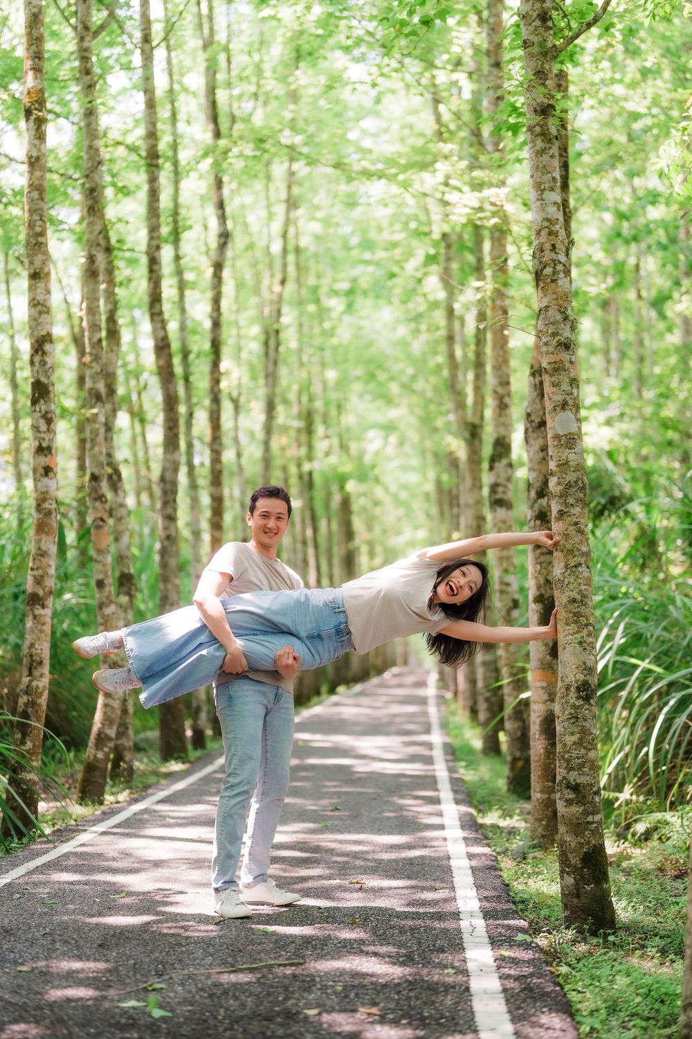 A joyful couple poses playfully on a tree-lined path, man lifting woman horizontally as she extends her arms wide.