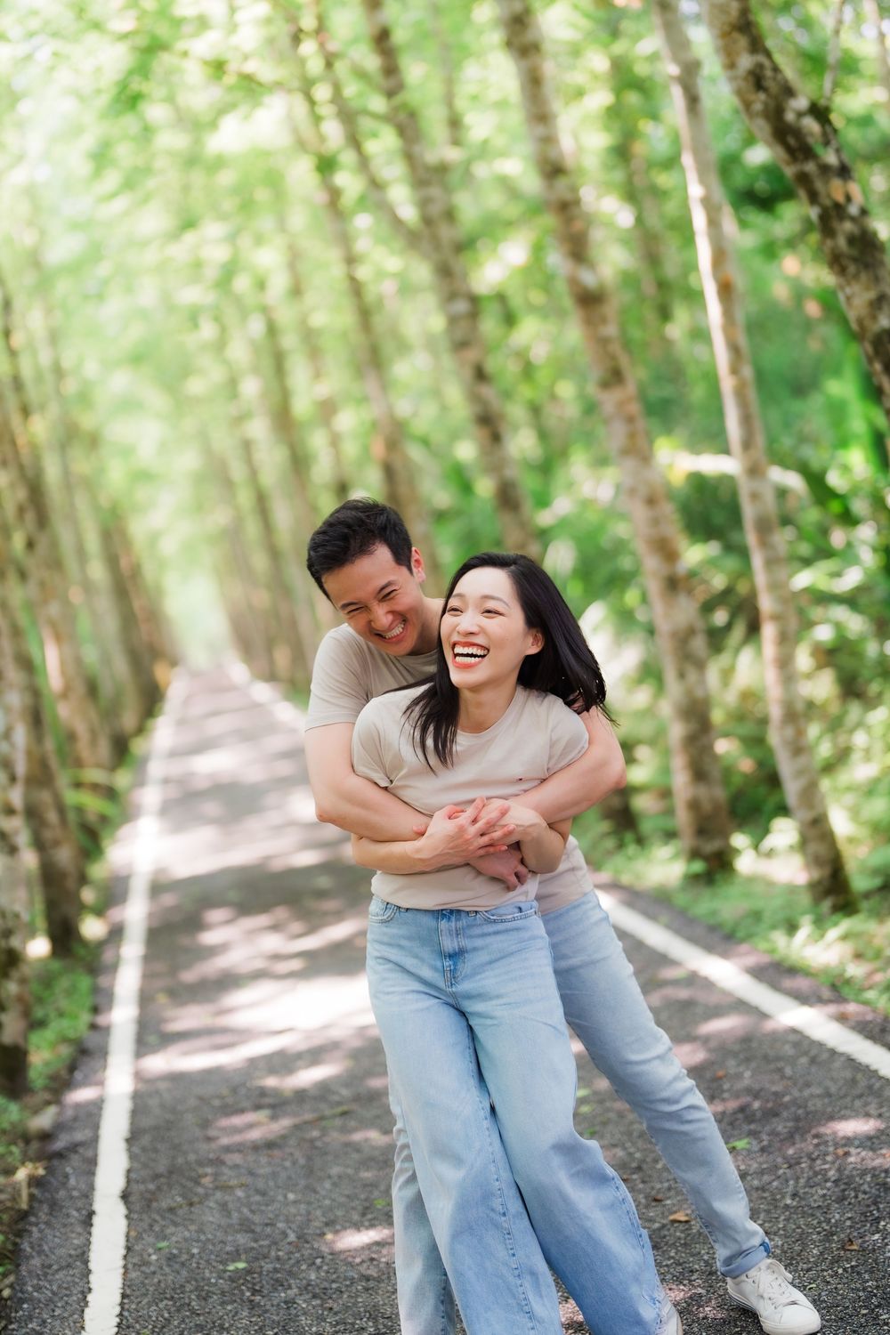 Happy Asian couple embracing and laughing together on a tree-lined road surrounded by lush green forest.