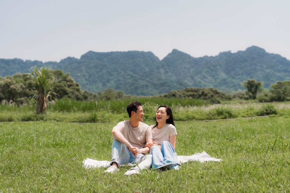 Happy Asian couple sitting together on a blanket in a green meadow with mountain views.