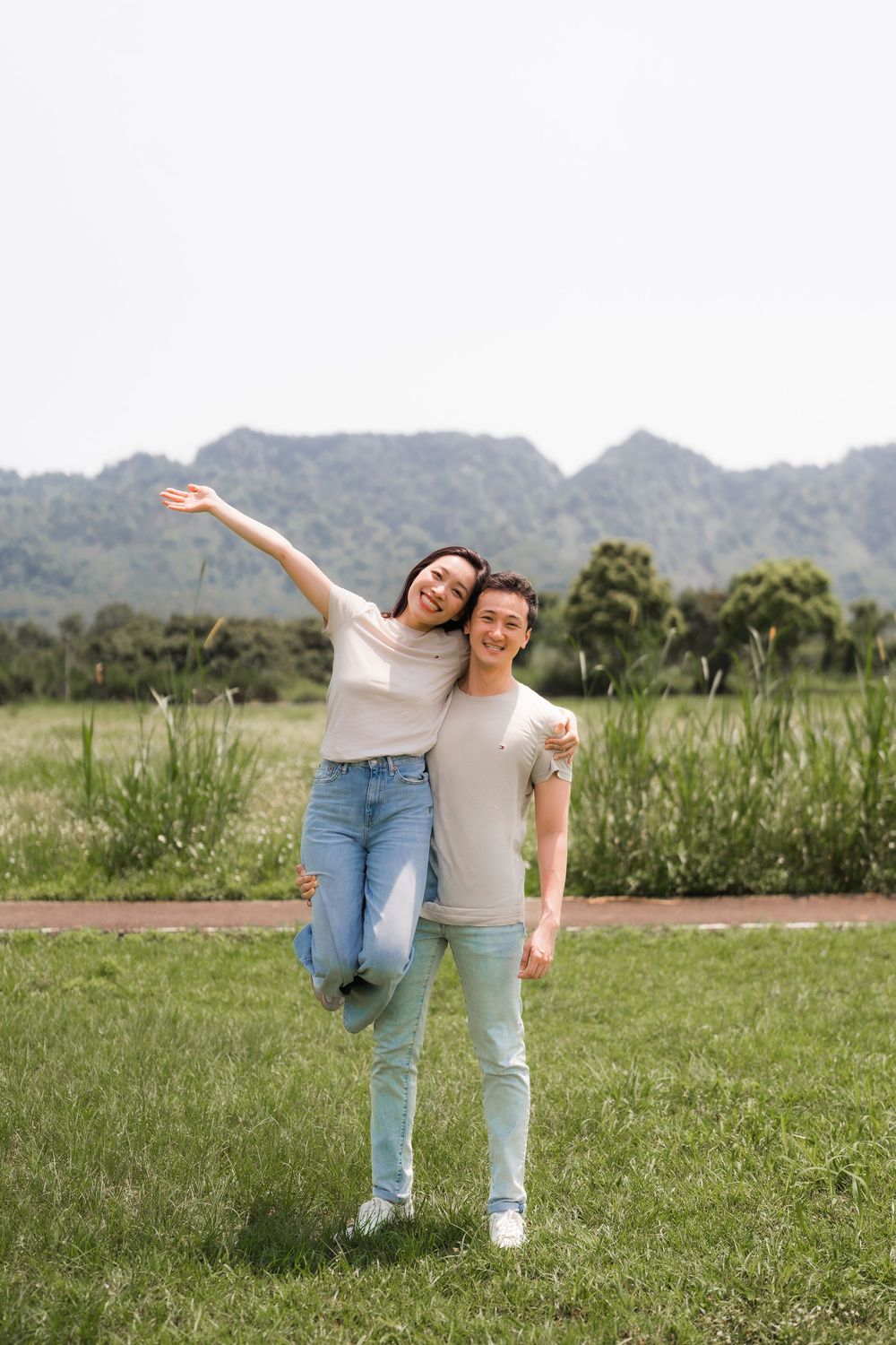 Joyful Asian couple posing together in a green field with mountains, woman raising her arm freely.