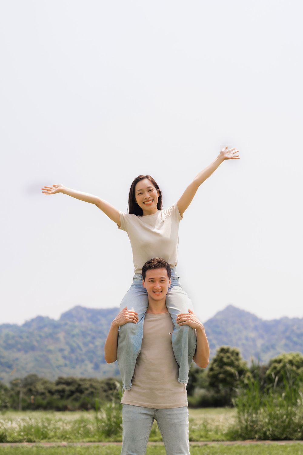 Happy couple outdoors, woman sitting on mans shoulders with arms raised triumphantly against mountain backdrop.
