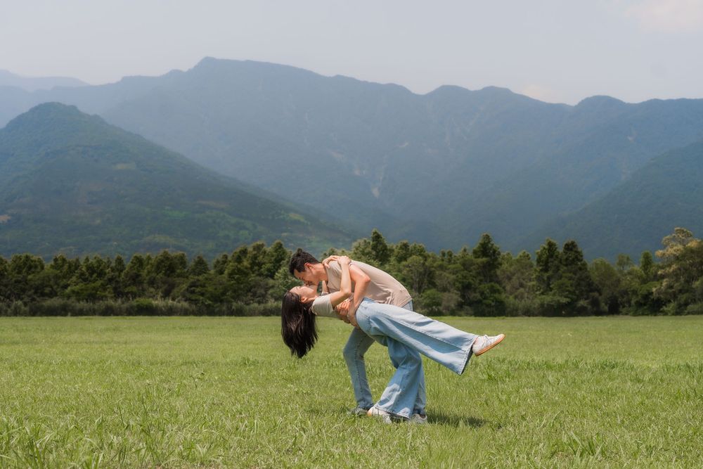 Couple sharing a romantic dip kiss in a lush green meadow with stunning mountain backdrop.