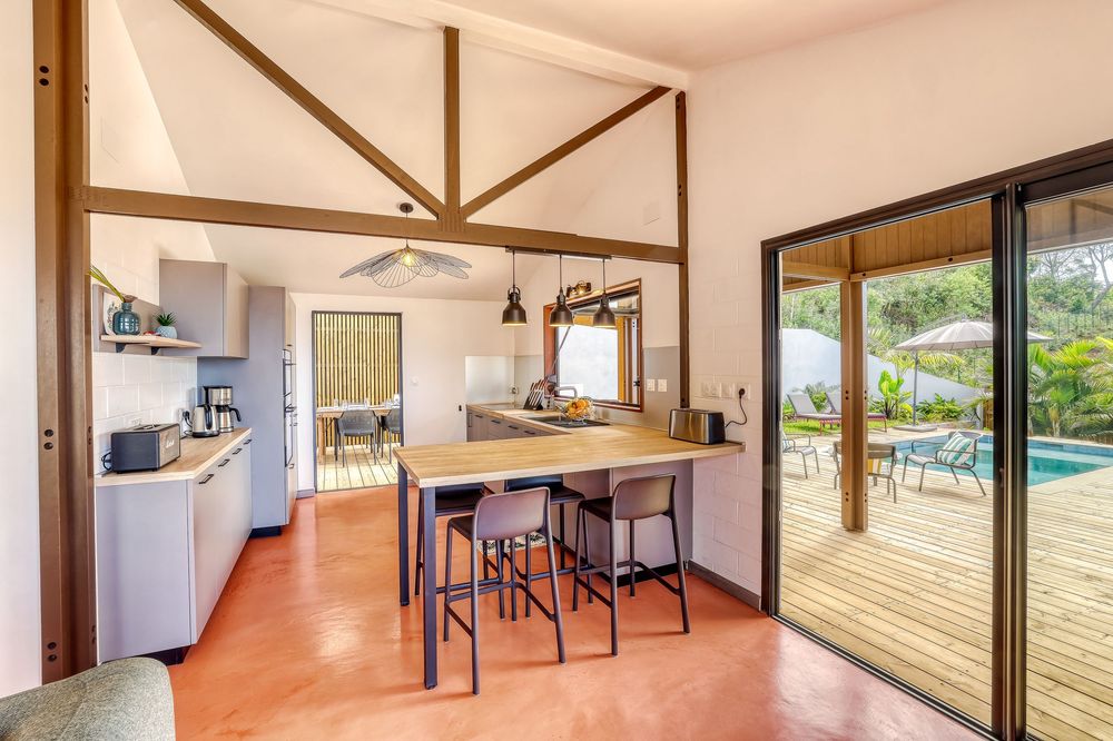 Modern open-plan kitchen and dining area with high vaulted ceilings, bar stools, and sliding doors leading to a sunny pool deck.