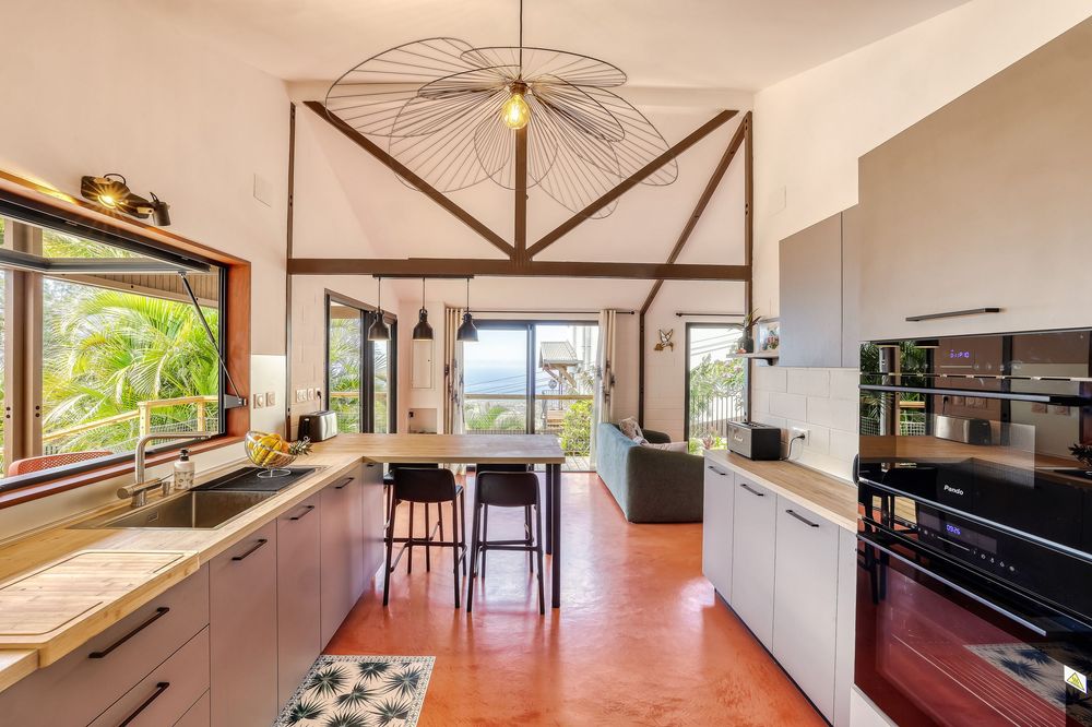 Modern open-plan kitchen with vaulted ceilings, gray cabinets, terracotta floors, and tropical garden views through large windows.