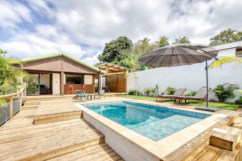 Tropical villa with a raised swimming pool on a wooden deck, lounge chairs, umbrella, and lush greenery.