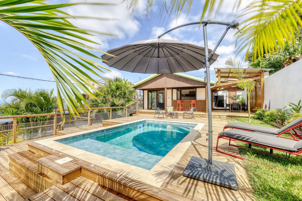 Tropical villa with a sparkling pool, wooden deck, lounge chairs, and a large umbrella surrounded by lush greenery.