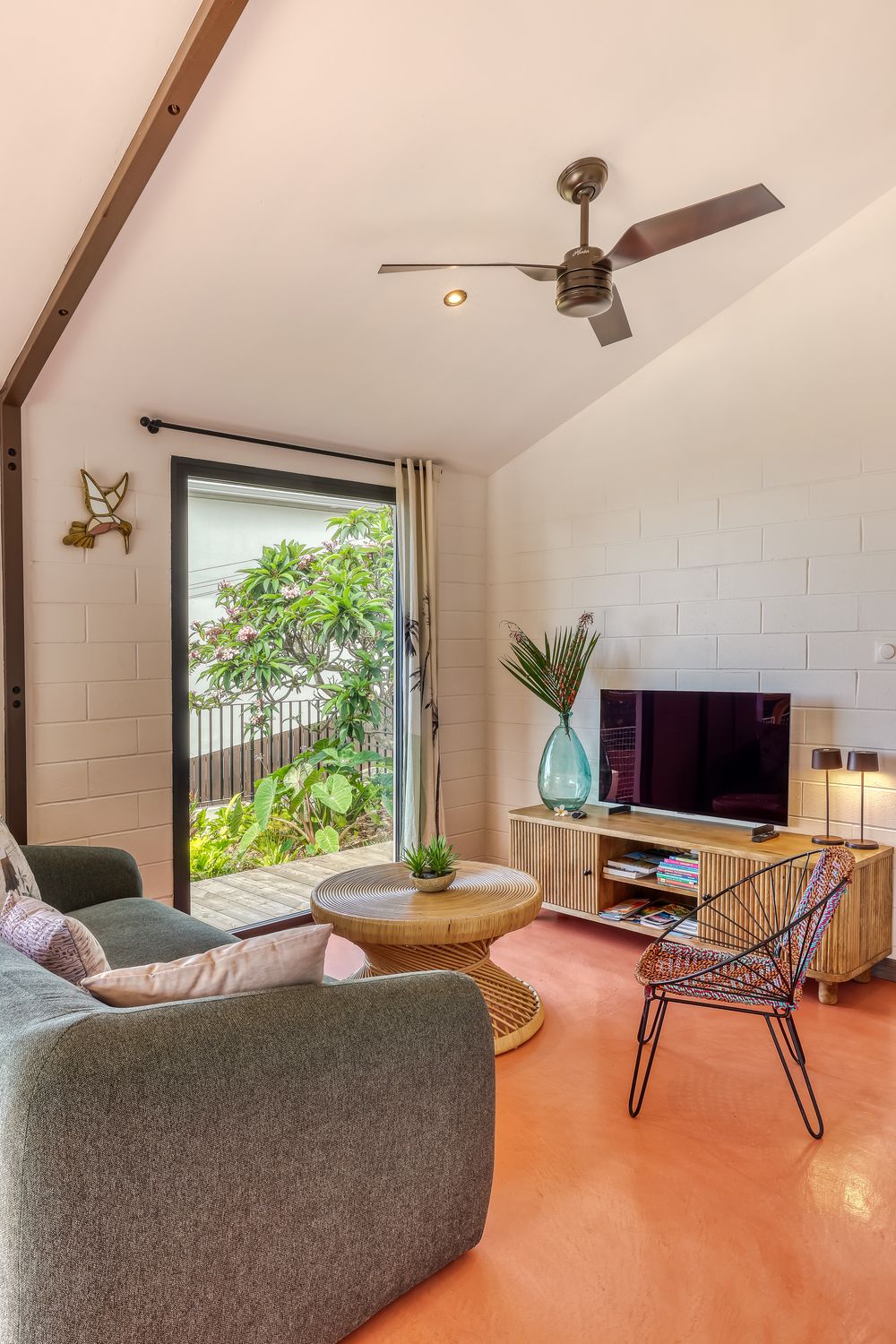 Cozy living room with vaulted ceiling, ceiling fan, gray sofa, wooden TV stand, and open door to lush garden.