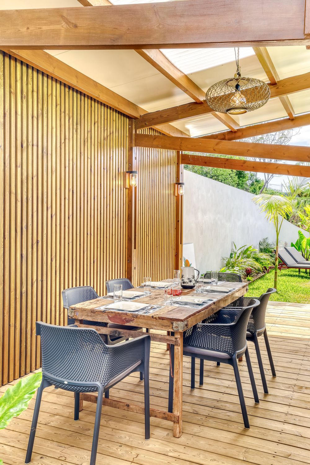 Outdoor covered dining area with wooden deck, slatted walls, rustic table, gray chairs, and pendant lighting.