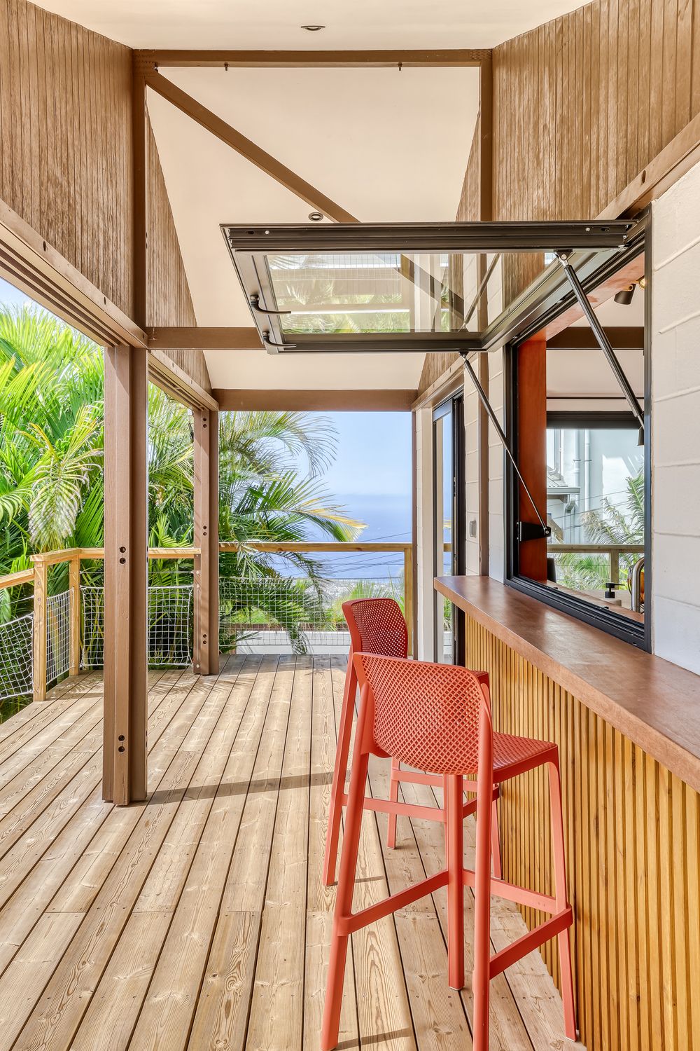 Tropical deck with red bar stools, wood countertop, bifold window, and ocean view surrounded by lush greenery.