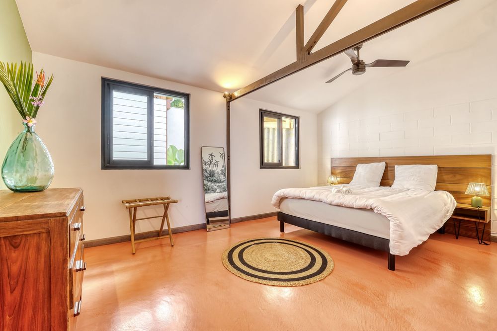 Spacious bedroom with wooden headboard, white bedding, ceiling fan, hardwood floors, and tropical plant by bright windows.
