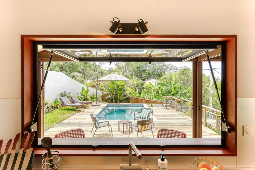 View through an open kitchen window of a tropical pool deck with outdoor dining set and umbrella.