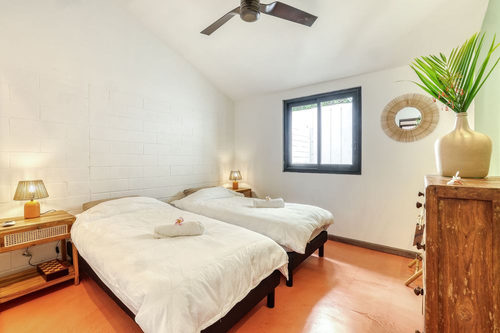 Twin bedroom with white walls, terracotta floors, ceiling fan, and natural light from a small window.