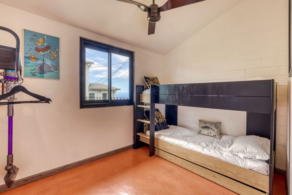 Compact bedroom with bunk bed, terracotta tile floor, blue-framed window, ceiling fan, and colorful wall art.