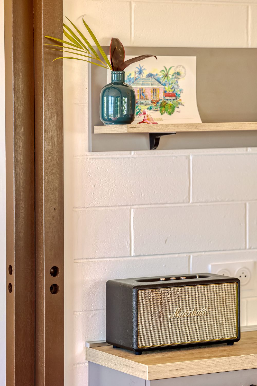 Marshall Stanmore speaker on wooden shelf beside white brick wall with blue vase and tropical artwork above.